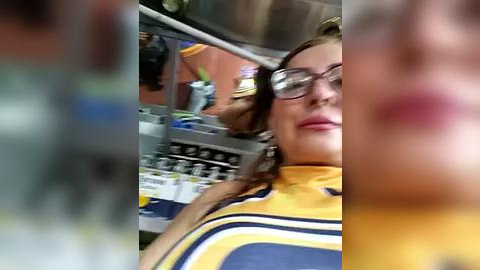 Media: A blurred video of a woman with glasses, wearing a yellow and blue striped t-shirt, and a red lipstick. The background shows a messy kitchen counter with multiple items.