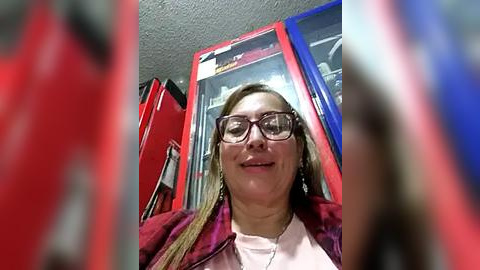 Media: A video of a smiling woman with glasses, wearing a pink top and purple jacket, standing in front of red and blue vending machines in a dimly lit room.