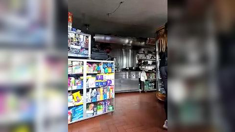 Media: Video of a well-lit, modern kitchen with stainless steel appliances, white countertops, and tiled floor. A person in a white shirt stands near the stove, and colorful boxed cereals are displayed on a metal shelf.