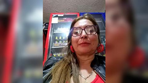 Media: Video of a middle-aged woman with glasses, wearing a black jacket over a red top, red headphones, and a necklace. She stands in front of a red and blue arcade machine with various game controllers.