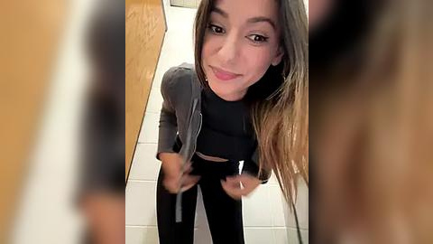 Media: Video of a young, light-skinned woman with long brown hair, wearing a black cropped top and high-waisted black pants, taking a selfie in a bathroom with tiled walls.