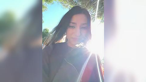 Media: Video of a young woman with long, dark hair, wearing a black jacket, standing outdoors with sunlight streaming through trees.