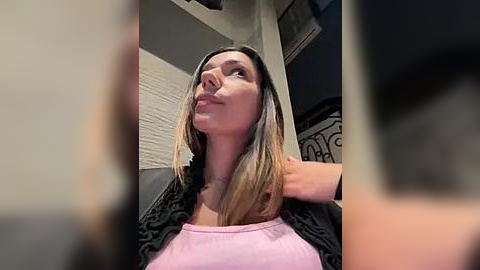 Media: A video of a woman with light skin and shoulder-length blonde hair, wearing a black lace cardigan over a pink tank top, standing indoors with a contemplative expression.