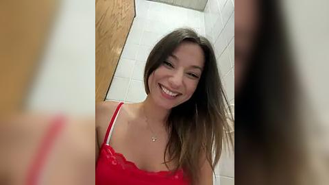 Media: Video of a smiling, young Asian woman with long brown hair, wearing a red lace bra, standing in a tiled bathroom with wooden door.