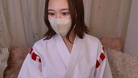 Media: Video of an Asian woman with shoulder-length brown hair, wearing a white kimono with red accents and a beige face mask, seated indoors with beige curtains and a soft, white furry rug in the background.