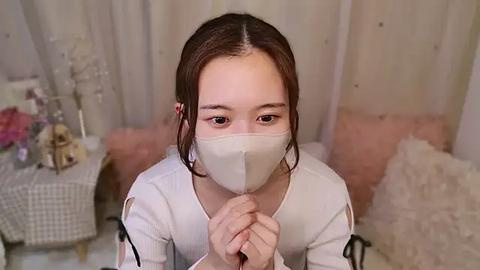 Media: Video of an Asian woman with brown hair and light skin wearing a white mask and long-sleeved top, kneeling with clasped hands in a cozy, softly lit room with pink and white decor.