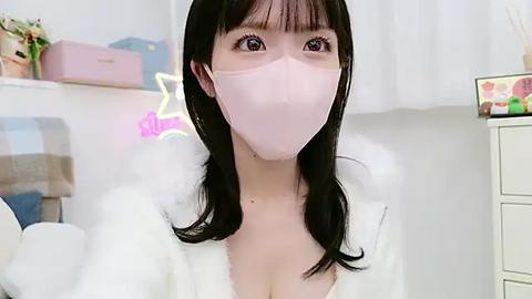 Media: Video of an Asian woman with long black hair, wearing a white robe and a pink surgical mask, standing in a bright, minimalist room with white walls and pastel decor.