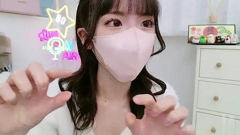 Media: Video of a young East Asian woman with long dark hair, wearing a white mask, adjusting her hair, in a bright, minimalistic room with pastel colors.