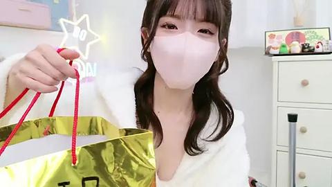 Media: Video of a young woman with fair skin, long brown hair, and bangs, wearing a white mask and white robe. She holds a gold-patterned shopping bag with red handles. Background features a white dresser and colorful toys.