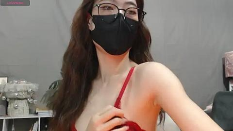 Media: A video of a young woman with long brown hair, wearing glasses and a black face mask, holding a red bra, in a dimly lit room with shelves in the background.