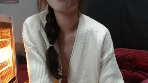 Media: Video of a young Asian woman with a braided ponytail, wearing a white ribbed robe, standing indoors near a bed with red bedding and a wicker lamp.