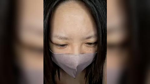 Media: Video of an Asian woman with straight black hair, wearing a light pink face mask, looking downward with a solemn expression. Background is blurred, focusing attention on her face.