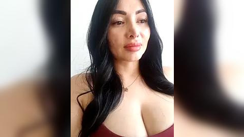 Media: Video of a Latina woman with long, black hair and fair skin, wearing a burgundy off-shoulder top that accentuates her large breasts. She has a neutral expression, standing against a plain, light-colored background.