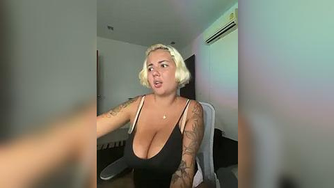 Media: Video of a curvy blonde woman with tattoos, wearing a low-cut black top, sitting on a white chair, looking surprised in a modern, minimalist room with an air conditioner.