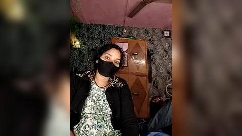 Media: Video of a woman with dark skin, wearing a black mask, black jacket, and a white floral blouse, seated in a dimly lit room with patterned wallpaper, wooden furniture, and a ceiling fan.