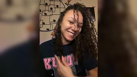 Media: A candid video of a joyful, curly-haired Black woman with a broad smile, wearing a \"KIMON\" t-shirt. She's in a cozy, dimly-lit room with a geometric-patterned wall decoration in the background.