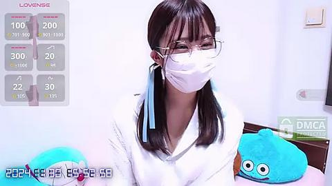 Media: A video of a young East Asian woman with straight black hair, wearing a white mask, glasses, and a white top, standing in a bedroom with blue plush toys.