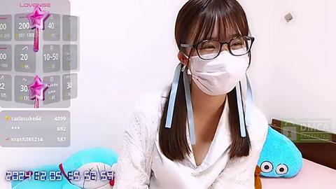 Media: A video of an East Asian woman with long straight brown hair, wearing glasses, a white mask, and a white sweater, sitting on a pink table. The background features a chart, plush toys, and a box labeled \"Dining.\