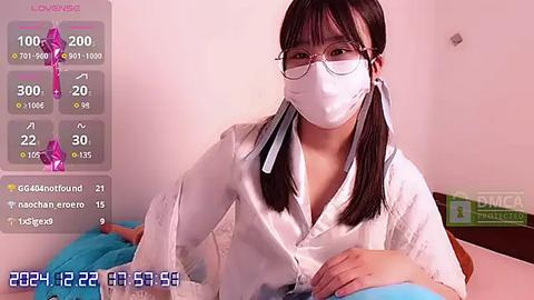 Media: Video of a young Asian woman with glasses and a white mask, wearing a white robe, sitting on a bed in a pink room.