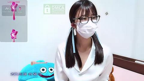 Media: Video of an Asian woman with straight black hair in pigtails, wearing black-rimmed glasses, white mask, and white shirt, sitting in a bright, minimalistic room with a colorful plush toy.