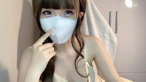 Media: A young Asian woman with long brown hair, wearing a white face mask, a white spaghetti strap top, and a towel, stands in a light-colored room, pointing at her face with her index finger.
