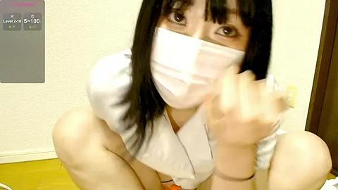 Media: Video of an Asian woman with long black hair, wearing a white surgical mask, crouching, pointing at the camera. She's in a room with a beige wall and wooden floor.