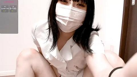 Media: Video of an East Asian woman with long black hair, wearing a white surgical mask, white button-up shirt, and white panties, sitting on the floor in a clinical setting, capturing a casual, intimate moment.