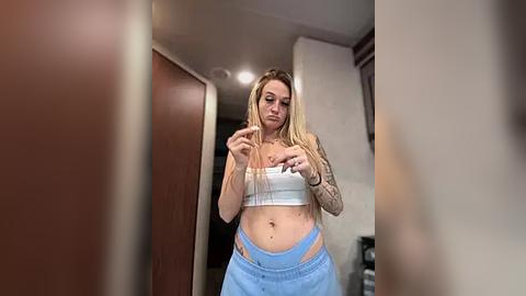 Media: A video of a blonde woman with tattoos, wearing a white crop top and blue pants, taking a selfie in a dimly lit hallway.