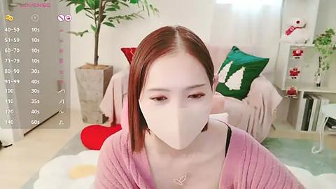 Media: Video of a young woman with fair skin, straight red hair, and a pink knit cardigan, wearing a white mask, in a cozy, softly lit room with plants and decorative pillows.