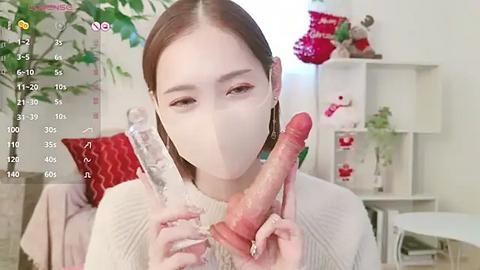 Media: Video of an East Asian woman with fair skin, brown hair, and wearing a white mask, holding two large, realistic, erect pink dildos, in a modern, minimalist room with white walls and festive decorations.