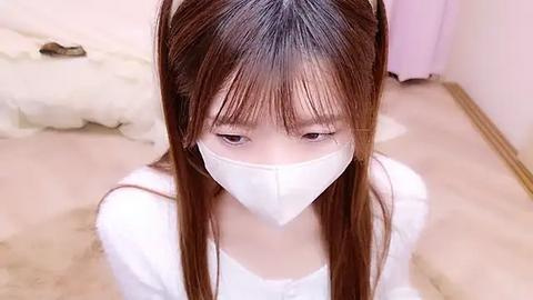 Media: A video of a young Asian woman with long, straight brown hair, wearing a white face mask and a white top, kneeling on a beige carpeted floor in a softly lit, minimalist room.