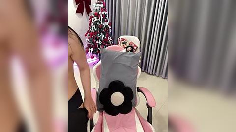 Media: Video of a young woman with light skin, sitting in a pink gaming chair, wearing a gray hoodie, and a plush toy on her head. Background features a Christmas tree and silver curtains.
