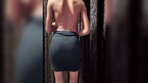 Media: Video of a slender, light-skinned woman from behind, topless, wearing a tight black skirt, standing against dark, patterned curtains. The image is slightly blurred, emphasizing her hourglass figure and the contrast between her skin and the fabric.