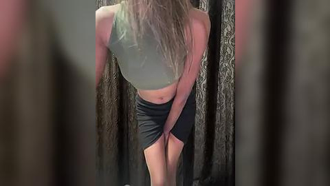 Media: Video of a slender, blonde woman with long hair, wearing a green crop top and black skirt, standing against dark, patterned curtains in a dimly lit room.