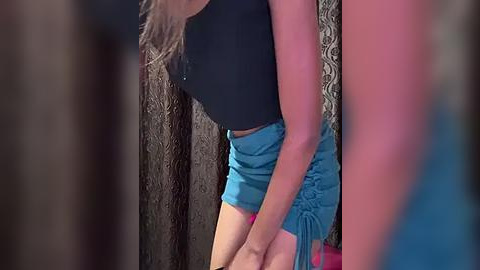 Media: Video of a young woman with light skin and long brown hair, wearing a black crop top and blue shorts with a ruffled waistband, leaning against a textured brown wall.