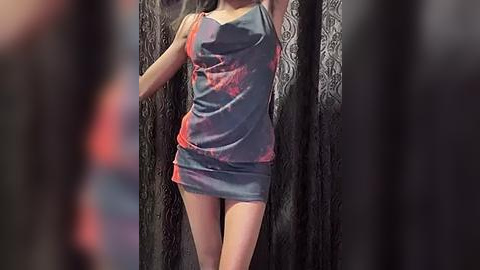 Media: Video of a slender, light-skinned woman with long, dark hair wearing a tight, grey mini dress with red and black patterns, standing against black, textured curtains.