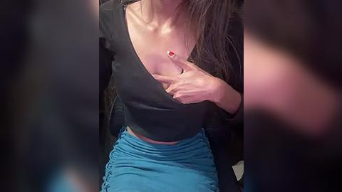 Media: Video of a woman with long, dark hair, wearing a black top and blue skirt, lifting her shirt to expose her breast, with blurred background.