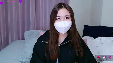 Media: Video of a young Asian woman with long brown hair, wearing a white face mask, black hoodie, and sitting on a bed with white and pink sheets, lavender curtains in the background.