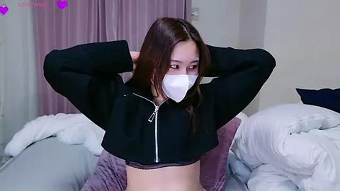 Media: Video of an Asian woman with long brown hair, wearing a black cropped hoodie, white mask, and white bra, adjusting her mask. Background shows a messy bed with white and purple bedding.