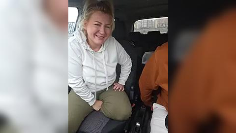Media: Video of a smiling, fair-skinned blonde woman in a white hoodie and olive green pants, sitting in a car next to an orange-jacketed man.