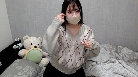 Media: Video of a woman with short black hair, wearing a face mask, beige sweater, and black pants, adjusting her glasses, surrounded by a gray, floral-patterned bedspread.