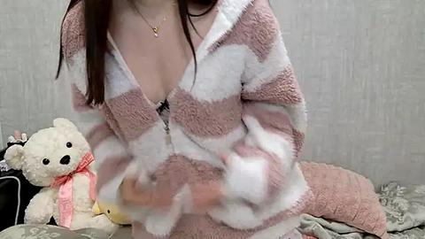 Media: Video of a light-skinned woman in a pink and white striped fuzzy pajama top, showcasing her cleavage. Background features a teddy bear and a patterned gray wall.