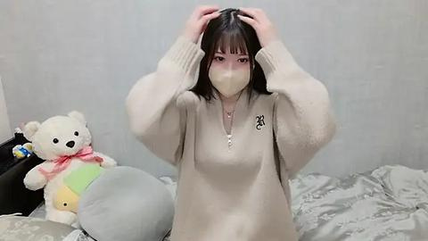 Media: A video of an East Asian woman with pale skin, wearing a beige sweater, face mask, and holding her long, dark hair. She sits on a bed with a stuffed animal, a teddy bear, and a pillow.