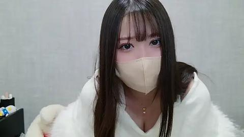 Media: Video of an East Asian woman with long, straight black hair, wearing a beige face mask and white fluffy robe, sitting indoors against a plain gray background.