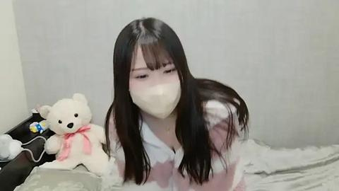 Media: Video of an Asian woman with long black hair, wearing a white mask, pink robe, and lying on a bed with a teddy bear.