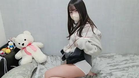 Media: Video of an Asian woman with long black hair, wearing a white jacket, white shorts, and a surgical mask, sitting on a bed with a stuffed teddy bear and colorful toys in the background.