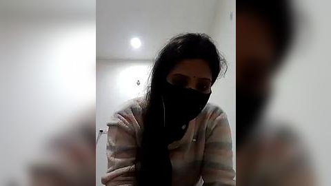 Media: Video of a woman with long dark hair, wearing a black face mask and a striped sweater, standing in a dimly lit room with a blurred background.