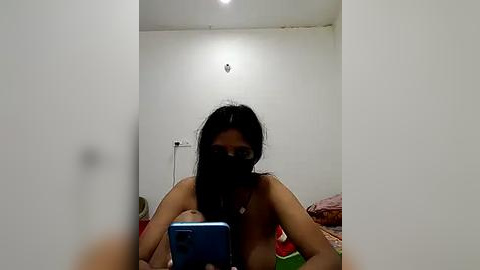 Media: A video of a topless South Asian woman with medium skin tone, long black hair, and dark eyes, taking a selfie in a dimly lit bedroom with white walls, a bed with red and green sheets, and a light fixture.