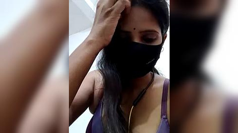 Media: Video of a woman with long black hair, wearing a black mask and purple top, adjusting her hair, blurred background.