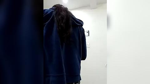 Media: Video of a person with long, dark hair, wearing a blue hoodie, walking in a dimly lit hallway with white walls and a single light bulb.